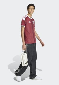 Vinröd sporttröja med vita detaljer och zigzagmönster, tillsammans med mörka, baggy jeans och vita sneakers, håller en vit väska.