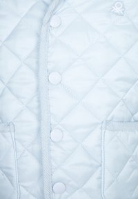 Světlomodrá prošívaná bunda s diamantovým vzorem, hladkou texturou a bílými plastovými knoflíky, s dekorativním logem a dvěma předními kapsami.