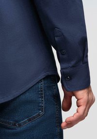 Homme portant une chemise à manches longues bleu foncé avec poignet boutonné et jeans en denim bleu, main détendue sur le côté, fond gris clair.
