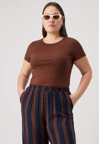Rjava rebrasta majica s kratkimi rokavi, v kombinaciji z mornarsko in rjavo črtastimi hlačami. Model nosi bele mačje očala in ima sproščen položaj.