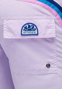 Pantaloni lilla con tasca a pattina quadrata, caratterizzati da un logo "SUNDEK" blu e un bottone circolare decorativo. Accenti a righe in rosa e blu.