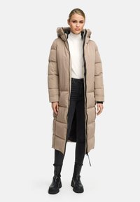 Giacca imbottita beige con cappuccio, chiusura frontale a zip e spacchi laterali. Tessuto liscio, motivo trapuntato, indossata sopra un dolcevita bianco e jeans scuri.