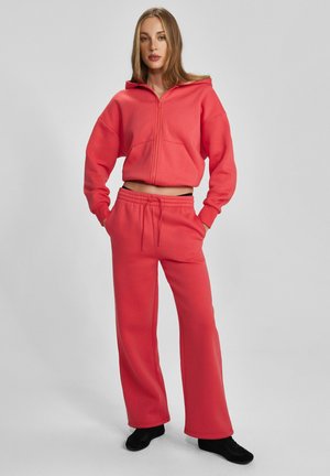 Kobieta stoi z rękami w kieszeniach, ubrana w koralowo-różową bluzę z kapturem oraz pasujące szerokie dresowe spodnie, na szarym tle, ma na nogach czarne buty.