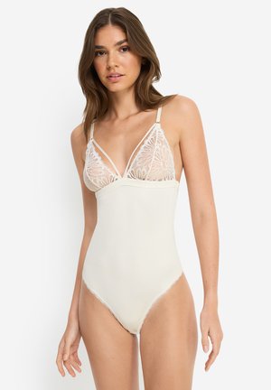 Junge Frau trägt weißen Spitzen- und Netz-Body mit floralen Mustern und verstellbaren Trägern, steht vor einfachem hellem Hintergrund.