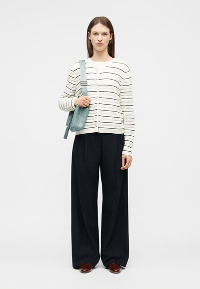 Γυναίκα που φοράει λευκή ριγέ πλεκτή ζακέτα, μαύρο παντελόνι με φαρδιά πόδια, καφέ παπούτσια και κρατάει ένα ανοιχτό μπλε τσαντάκι ώμου.