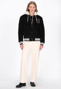 Fekete steppelt bomber dzseki fehér műszőrme kapucnival, "D" betűs díszítéssel és csíkos mandzsettákkal, bézs színű bőszárú nadrág fölött viselve.