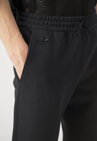 Pantaloni sportivi neri con elastico in vita e coulisse. Presentano tasche laterali e logo ricamato tono su tono sulla parte anteriore. Tessuto liscio e elasticizzato.