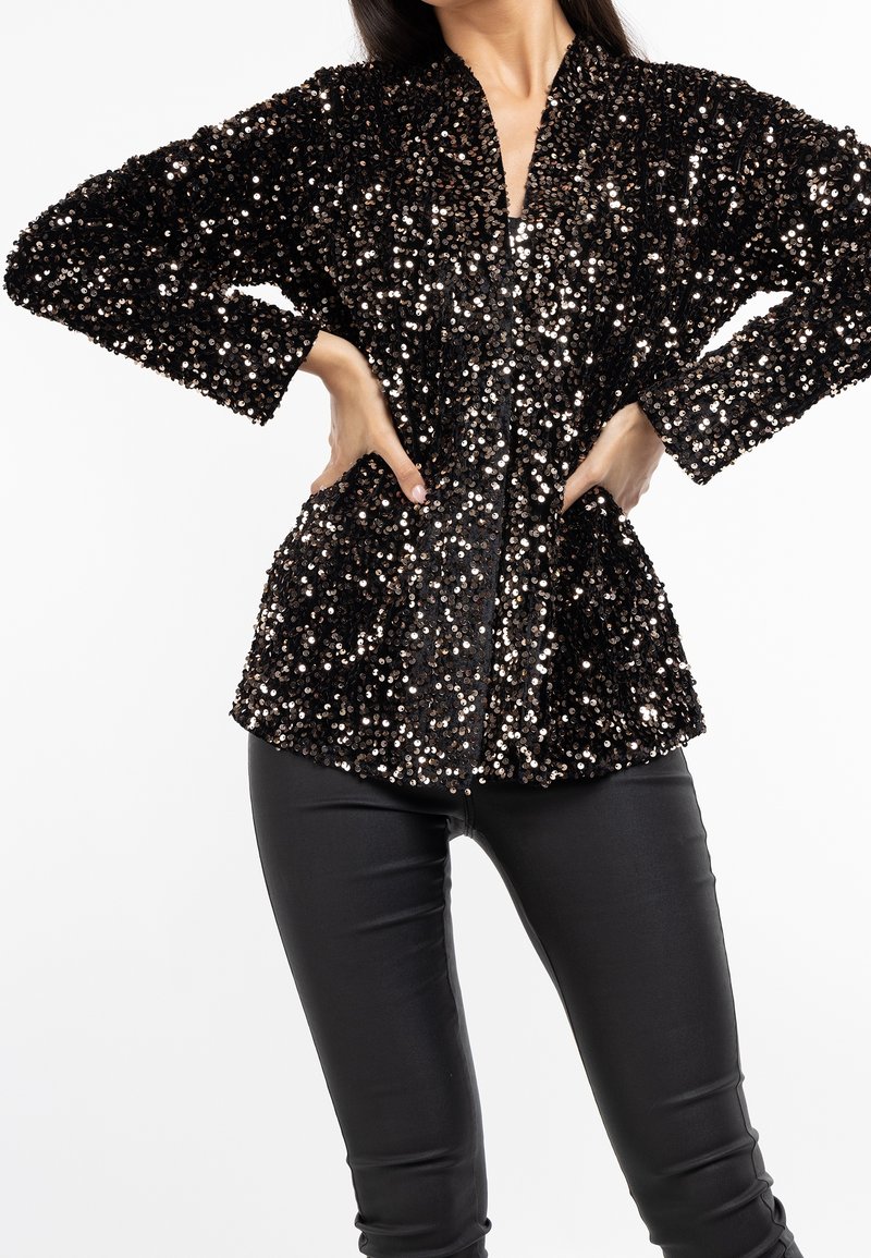 Femme portant un blazer noir à manches longues pailleté, les mains sur les hanches et un pantalon en cuir noir moulant, sur un fond blanc uni.