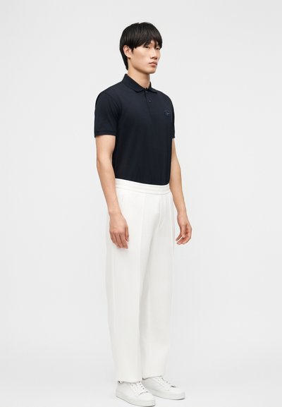 Νεαρός άνδρας που στέκεται, φοράει μαύρο πόλο με κοντά μανίκια, λευκό παντελόνι με φαρδιά πόδια και λευκά αθλητικά παπούτσια, μπροστά σε απλό φόντο.