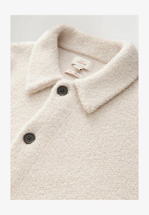 Chaqueta de forro polar crema con un cuello tipo camisa, que presenta dos botones negros y una superficie suave y texturizada. Etiquetada "NEXT SIGNATURE."