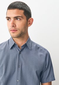 Chemise à manches courtes, boutonnée, en tissu bleu à motifs à carreaux, col pointu et boutons foncés. Texture lisse et coupe décontractée.