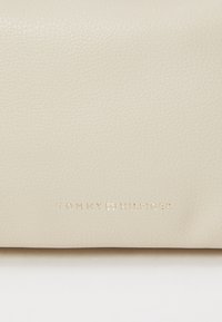 Pouch od krem kože s teksturiranom površinom; sadrži utisnuti logotip "TOMMY HILFIGER" u zlatnoj boji, minimalistički dizajn.