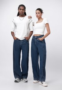 Två personer står sida vid sida klädda i avslappnade kläder. Båda bär löst sittande, mörka denimjeans med kontrasterande sidostrecks. Topparna är vita, en i t-shirt med kort ärm och den andra i en figurnära topp med fyrkantig halsringning.