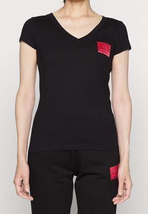 Žena oblečená v černém tričku s krátkými rukávy a výstřihem do V a černých kalhotách, obojí s červeným obdélníkovým logem na levé straně.