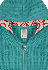 Tiefgrüne Zip-Hoodie aus strukturiertem Stoff, mit einem Innenfutter in rot-weißen Löwenmustern und einer gemütlichen Kapuze.