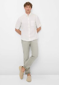 Camicia bianca in lino con maniche arrotolate, pantaloni slim-fit verde chiaro e scarpe beige senza lacci. Design semplice con taschino sul petto.