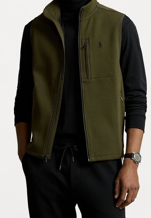 Olijfgroene zip-up vest met hoge kraag, verstelbare zoom en ritszak op de borst. G gedragen over een zwarte langemouwen top.