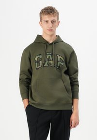 Fiatalember olíva zöld kapucnis felsőben, amelyen terepmintás "GAP" felirat látható, fekete nadrágban áll, egyszínű fehér háttér előtt.