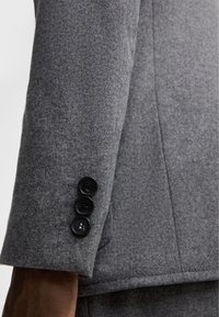 Ärmel eines grauen Wollblazers mit glatter Textur, drei schwarzen Knöpfen am Bund und taillierter Passform.