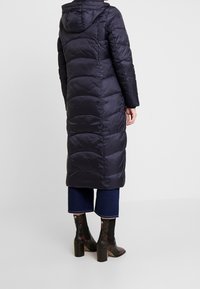 Dlouhý tmavě modrý péřový kabát s kapucí, s horizontálním prošíváním a hladkou, jemnou texturou. Kombinován s krátkými džínami a botami na podpatku.
