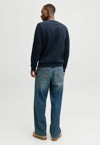 Marineblauw sweatshirt met geribbelde mouwen en onderkant, gecombineerd met losse, vervaagde blauwe jeans en bruine schoenen, bekeken vanuit de achterkant.