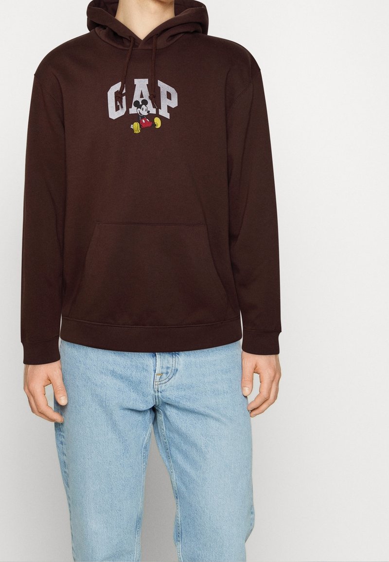 Personne portant un sweat à capuche marron foncé avec « GAP » et un motif de Mickey Mouse, associé à un jean bleu clair sur un fond uni.