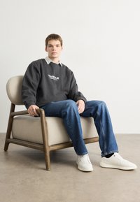 Šedá oversized mikina s bílým logem, nošená přes košili s límečkem; modré džíny; bílé tenisky; sedící na světlé židli.