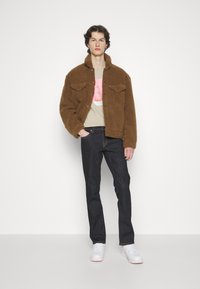 Bruine schapenvachtjas met twee borstzakken, gedragen over een lichtbeige T-shirt met een grafische print, gecombineerd met donkere spijkerbroek en witte sneakers.