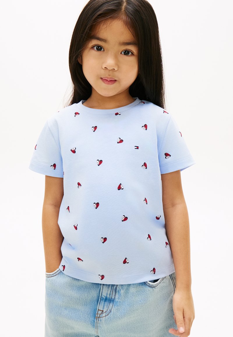 Νεαρή κοπέλα με μακριά σκούρα μαλλιά που φοράει ανοιχτό μπλε πουκάμισο με μικρά κόκκινα και λευκά σχέδια σε σχήμα καρδιάς και ανοιχτόχρωμο τζιν παντελόνι.