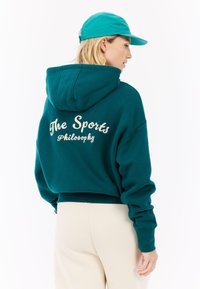 Tyrkysová skrátená mikina s bielym textom "The Sports Philosophy" na zadnej strane. Má predné vrecko a veľkú kapucňu. Kombinovaná so svetlo béžovými nohavicami.