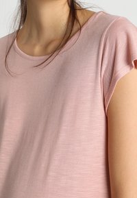 Light pink short-sleeved top with a round neckline. Soft, textured fabric with a relaxed fit. Subtle sheen and slight draping at the shoulders.