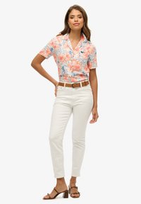 Koszula z krótkim rękawem w kwiatowy wzór w kolorze koralowym i białym z niebieskimi akcentami, zestawiona z białymi dżinsami i brązowym paskiem; modelka ma na sobie brązowe sandały na obcasie.