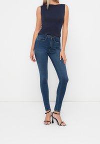 Top sin mangas color navy combinado con jeans ajustados de un azul oscuro. Diseño de cintura alta, tejido suave, con sandalias de tiras en blanco y negro de punta abierta.