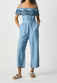 Kobieta ubrana w jasnoniebieski kombinezon z odkrytymi ramionami, z koronkowymi detalami w kwiaty oraz białymi sandałami na klamrze, z rękami w kieszeniach, stojąca na białym tle.
