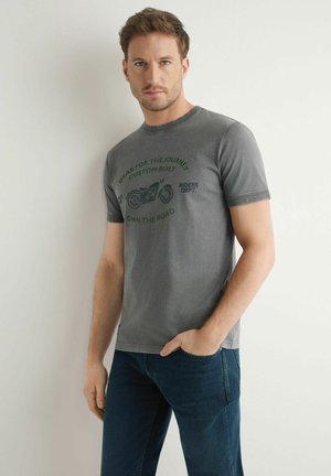 Mężczyzna o jasnobrązowych włosach, ubrany w szarą koszulkę z motocyklowym nadrukiem i niebieskie jeansy, stojący oparty o białą ścianę z jedną ręką w kieszeni.