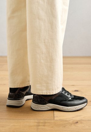 Sneaker sportivi neri con dettagli texturizzati e suole in gomma grigia, abbinati a pantaloni larghi color crema su un pavimento in legno.