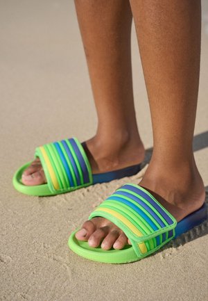 Pieds portant des sandales slides rayées vertes, bleues et jaunes, debout sur un sol sablonneux.