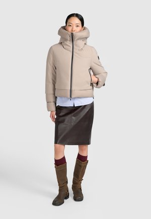 Femme portant une veste matelassée beige à col haut, une jupe en cuir foncé, des chaussettes bordeaux et des bottes marron hautes jusqu'aux genoux, debout devant un fond uni.