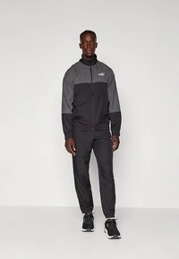 Άνδρας ντυμένος με γκρι και μαύρο Puma φόρεμα γυμναστικής και μαύρα αθλητικά παπούτσια μπροστά σε απλή αχνή φωτεινή φόντο.