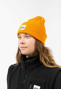 Orange ribbed beanie with a tag featuring black text, worn by a person in a black fleece vest, against a white background.