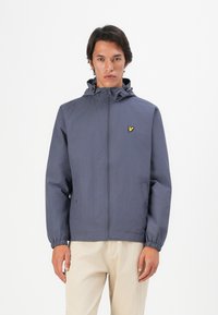 Homme portant une veste à capuche zippée bleu marine avec un logo d'oiseau jaune, et un pantalon beige, se tenant contre un fond blanc.