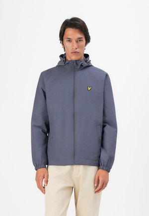 Man in een marineblauwe capuchon met rits en een gele vogel logo, met beige broek, staand tegen een witte achtergrond.