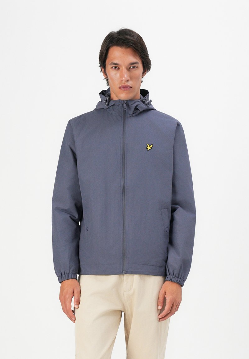 Homme portant une veste à capuche zippée bleu marine avec un logo d'oiseau jaune, et un pantalon beige, se tenant contre un fond blanc.
