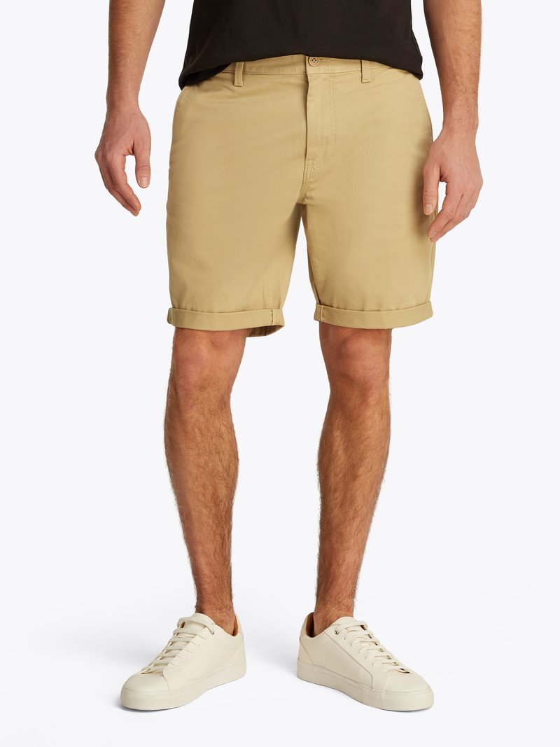 Calções bege com uma textura suave, com bainhas dobradas e passantes para cinto, combinados com ténis brancos. Design simples, adequado para uso casual.