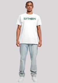 Hvit bomulls t-skjorte med et grønt "SLYTHERIN"-motiv. Kombinert med lyseblå jeans og hvite sneakers, står foran en grå bakgrunn.