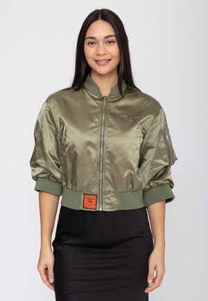 Donna sorridente, indossa una giacca bomber color oliva in satin con polsini a coste e una gonna nera, in piedi davanti a uno sfondo neutro.