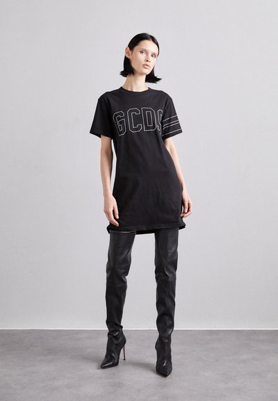 Γυναίκα που στέκεται μπροστά σε μια απλή τοιχώμα, φορώντας μια μαύρη υπερμεγέθη φόρμα GCDS και μαύρες δερμάτινες μπότες στιλέτο μέχρι το μηρό.