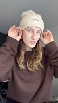 Bež beanie od rebrastog pletenja s kvadratnom etiketom na kojoj piše "MSCH COPENHAGEN." Model podešava beanie dok nosi tamnosmeđi pulover.