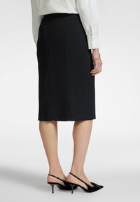 Zwarte kokerrok met een strakke pasvorm, gladde textuur, knielang en een achterkant met split, gecombineerd met zwarte slingback hakken.