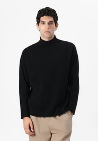 Maglione a collo alto di colore nero in tessuto morbido, con una vestibilità rilassata e una texture a coste, abbinato a pantaloni beige chiaro.